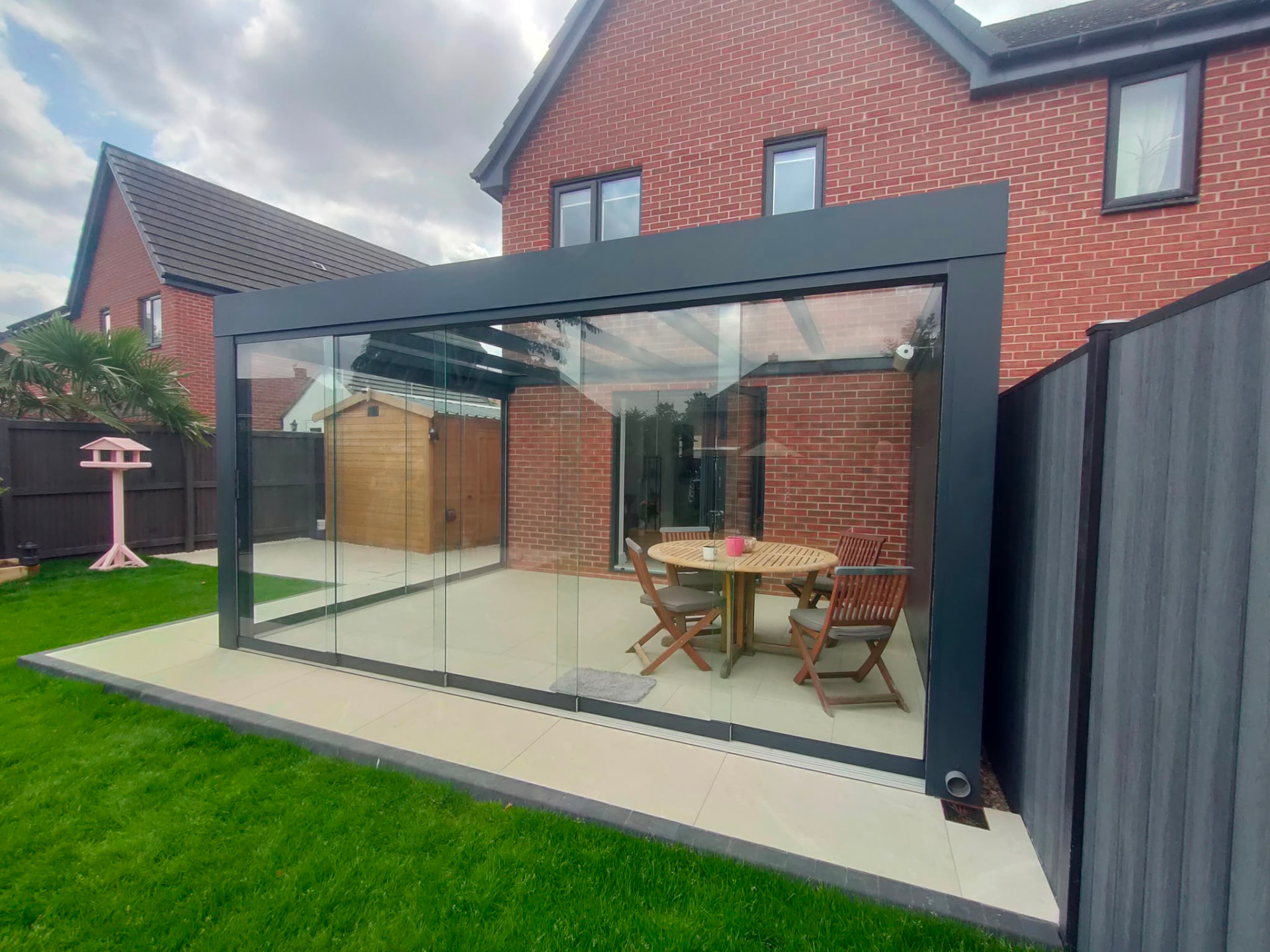 Glass room enclosing a patio dining area beside a red brick home